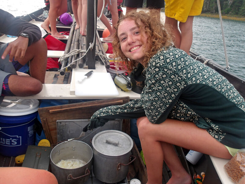 A young woman with curly hair smiles at the camera while crouching in a boat. She is surrounded by cooking equipment, including pots and a cutting board. Other people are visible in the background, suggesting a group outing on the water. The atmosphere appears casual and friendly.